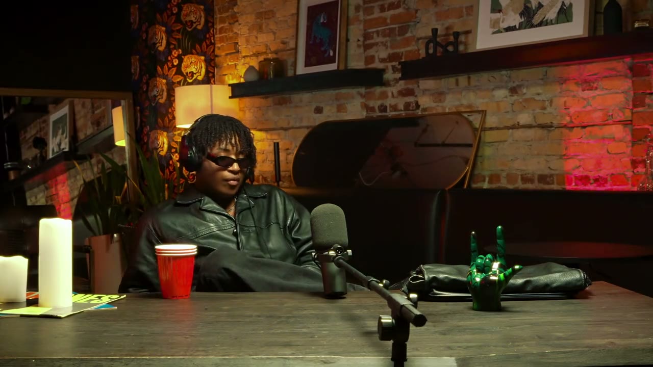 The room. Damian B seated at a dressed table under warm lamps, red wash on the brick behind him.