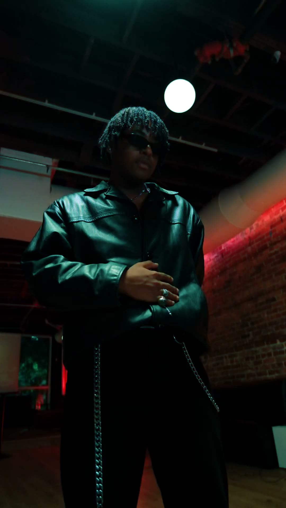 Damian B full length under red light and one hard white pendant, exposed brick behind him.